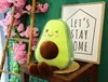 A plush avocado toy sits on a wooden surface next to a decorative ladder with pink flowers and a framed picture with text. Produktbild-Vorschau