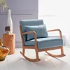 A blue fabric rocking chair with wooden arms and legs, placed in a well-lit room with a window in the background. A decorative vase and a wooden stump side table are also visible. Produktbild-Vorschau