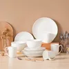 A collection of white ceramic dinnerware, including plates, bowls, and mugs, arranged on a wooden surface against a light brown background. Náhled obrázku produktu