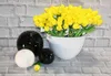A white pot of yellow tulips sits on a gray brick surface, next to three decorative spheres (one white, two black). Produktbild-Vorschau