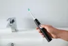 A hand holding a black electric toothbrush over a white sink with a chrome faucet. Voir les images du produit