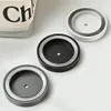 Three circular hair dryer holders are arranged on a light gray surface, with a partially visible glass container with text on it in the background. Produktbild-Vorschau