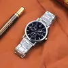 A silver watch with a black face is placed on a brown surface next to a pen. Produktbild-Vorschau