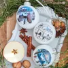 Christmas ornaments with winter village scenes, arranged with natural elements like pine branches, cinnamon sticks, star anise, an apple slice, and walnuts on a white tray. Náhled obrázku produktu