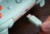 A hand is holding a mint green USB-C cable, about to plug it into a mint green and pink game controller. Produktbild-Vorschau