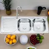 A stainless steel double kitchen sink with a faucet and a draining board, placed on a countertop. In the foreground, there are bowls of fruit and other items. Anteprima dell’immagine prodotto