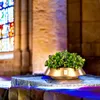 A decorative golden bowl filled with green plants sits on a stone surface inside a building with stained glass windows. Produktbild-Vorschau