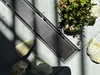 A shower drain with a brushed steel finish is placed on a tiled surface, surrounded by rocks and a small succulent plant. Sunlight casts shadows across the scene. Anteprima dell’immagine prodotto
