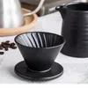 A black ceramic pour-over coffee dripper and a matching creamer pitcher are displayed on a textured surface, with coffee beans and a metallic kettle in the blurred background. Produktbild-Vorschau