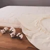A fitted sheet is draped over a mattress, with cotton branches on the wooden floor in the foreground. Produktbild-Vorschau