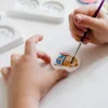 A child's hands are painting a small ceramic figurine of a rabbit. The rabbit is wearing a blue jacket and has pink ears. There are also white molds in the background. Produktbild-Vorschau
