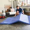 A man is setting up a blue folding gym mat in a living room with exercise equipment and furniture. Produktbild-Vorschau