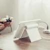 A person is playing a video game with a controller while a tablet is propped up on a stand on a wooden table. Náhľad obrázku produktu
