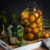 Two jars of preserved food, one with fruits and the other with vegetables and garlic, are placed on a wooden tray with cucumbers and dill. Voir les images du produit