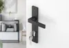 A black door handle with keys attached, mounted on a white door. The background shows a blurred interior scene with furniture and a plant. Produktbild-Vorschau