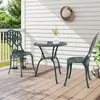 A green metal bistro set with two chairs and a table on a wooden deck, with a white wall and greenery in the background. Náhľad obrázku produktu