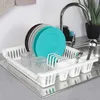 A white plastic dish drying rack is filled with clean dishes and glasses, placed next to a kitchen sink with a faucet and a plant in the background. Produktbild-Vorschau