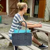 A woman sits at a wooden picnic table outdoors, holding a mug and looking to the side. A large grey felt tote bag with a bright blue interior sits on the bench next to her. Produktbild-Vorschau