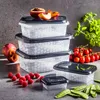 A set of clear food storage containers with black lids, arranged on a gray surface with fresh fruits and vegetables. Produktbild-Vorschau