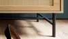 A close-up shot of the lower part of a wooden cabinet with a rattan front, supported by dark metal legs. A black high-heeled shoe is visible in the foreground on a wooden floor. Voir les images du produit