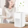 A woman is blurred in the background, sitting at a table, while a white air purifier is in the foreground on a wooden table. Green leaves are shown floating above the air purifier, suggesting clean air. Produktbild-Vorschau