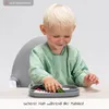 A young child wearing a green bib is sitting in a high chair and eating from a plate. Produktbild-Vorschau