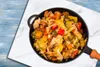 A close-up overhead view of a pan filled with cooked vegetables, placed on a marble surface with a blue wooden background. Anteprima dell’immagine prodotto