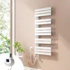 A white electric towel radiator is mounted on a light beige wall next to a window with greenery visible outside. A small white side table with a plant is also present. Voir les images du produit