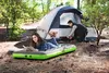 A young woman reads a book on an inflatable mattress in front of a tent, while a man sets up the tent. Podgląd zdjęcia produktu