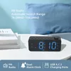 A digital alarm clock with FM radio and USB charging ports is displayed on a white table next to a stack of books, with a blue pillow in the background. Produktbild-Vorschau