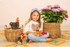 A young girl is sitting on a rug with stuffed chickens and a potted plant. Produktbild-Vorschau