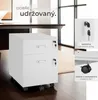 A white two-drawer filing cabinet on wheels, shown with close-ups of its lock and casters, set against a desk and a soft rug in an office-like setting. Náhled obrázku produktu