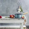 A teal saucepan with a wooden handle sits on a rustic wooden table next to plums and herbs. A bunch of garlic and rosemary hangs in the background. Náhled obrázku produktu