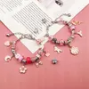 Two charm bracelets with various pink and silver charms laid out on a pink surface with a piece of paper with text in the background. Produktbild-Vorschau