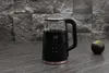 A black and glass electric kettle with a digital display sits on a dark countertop in front of a textured gray stone wall. A small potted plant is to the right of the kettle. Náhled obrázku produktu