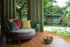 A cozy outdoor seating area with a round sofa, cushions, and a decorative bowl on a wooden deck, overlooking a garden with a modern house. Anteprima dell’immagine prodotto