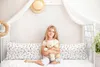 A young girl sits on a bed, hugging a teddy bear. The bed is covered with a floral-patterned sheet and has a canopy overhead. A straw hat hangs on the wall behind her. Produktbild-Vorschau