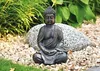 A Buddha statue sits in a garden setting, surrounded by gravel, rocks, and plants. Produktbild-Vorschau