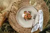 A plate of food is set on a woven mat with a napkin and fork, next to a glass of white wine. Anteprima dell’immagine prodotto