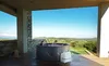 A couple relaxing in a hot tub on a wooden deck with a scenic view of rolling hills and the sky. Produktbild-Vorschau