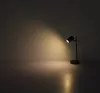 A black desk lamp is on a surface, casting a warm light on a plain wall. Produktbild-Vorschau