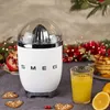 A white Smeg citrus juicer sits on a table next to a glass of orange juice, pancakes, and gingerbread cookies, with festive decorations in the background. Anteprima dell’immagine prodotto