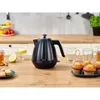 A kitchen counter scene with a black electric kettle, mugs, muffins, macarons, and fruit. Produktbild-Vorschau