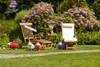 A garden scene with two deckchairs, a small table with drinks, and decorative sheep figures, set against a backdrop of blooming rhododendrons. Produktbild-Vorschau