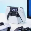 A white PlayStation PS5 controller alarm clock is displayed on a white surface, with a keyboard and a monitor in the background. Náhled obrázku produktu