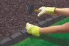 Hands wearing yellow gloves are using a rubber mallet to hammer a stake into the ground next to a garden border. Produktbild-Vorschau
