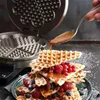 A stack of waffles with berries and caramel sauce being poured from a spoon, with a waffle maker in the background. Náhľad obrázku produktu