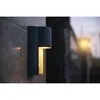 An outdoor wall light mounted on a textured stone wall, with a blurred background showing a fence and another light source. Anteprima dell’immagine prodotto