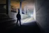 A person stands in a dimly lit concrete tunnel, illuminated by a bright light emanating from the tunnel's end. Produktbild-Vorschau