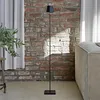 A black floor lamp stands in a room with a brick wall and a sofa. Produktbild-Vorschau